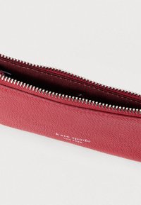 Červená kožená peňaženka so zipsom, ktorá má textúrovaný povrch, strieborný zips a na prednej strane reliéfne vyrazené logo. Vnútro je podšité textíliou.