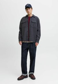 Chaqueta de camisa gris con bolsillos en el pecho, llevada sobre un suéter burdeos, combinada con jeans de mezclilla oscura y zapatos marrones con cordones.