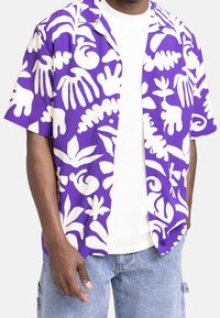 Man in paars en wit korter overhemd met abstracte bladeren en handafdruk over een witte T-shirt en lichtblauwe jeans.