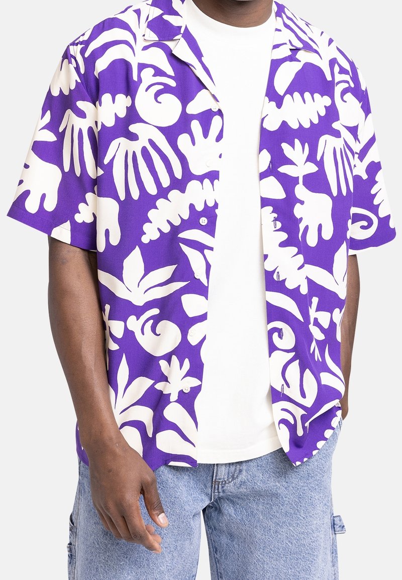 Man in paars en wit korter overhemd met abstracte bladeren en handafdruk over een witte T-shirt en lichtblauwe jeans.