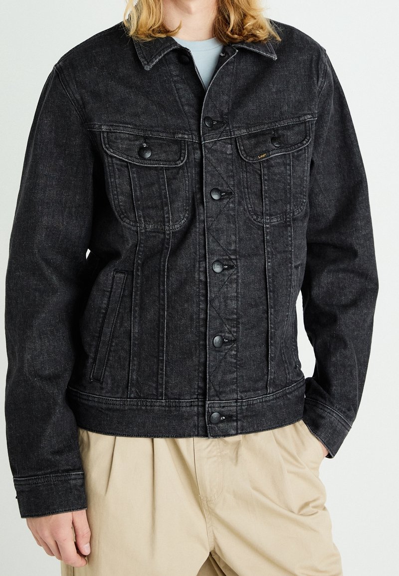 Personne portant une veste en jean noire avec des poches poitrine boutonnées et un pantalon beige, debout avec une main dans la poche et l'autre détendue.