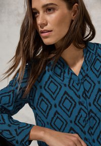 Camicia button-up teal con un motivo geometrico a diamante nero, dotata di colletto e maniche lunghe. Tessuto di consistenza liscia.