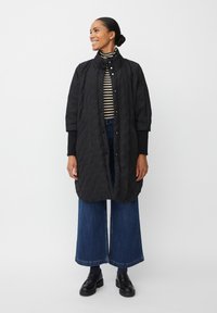 Svart kviltad kappa med hög krage, knappstängning och oversized passform. Bärs över en svart-vit randig polotröja och vida denimbyxor.