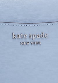 Ljusblått texturerat läder med silverfärgat logo "kate spade NEW YORK" tydligt placerad i mitten. Sömdetaljer längs kanterna.