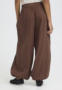 Pantalon marron ample avec taille et poignets élastiques porté avec des chaussures noires, vu de dos sur une personne debout.