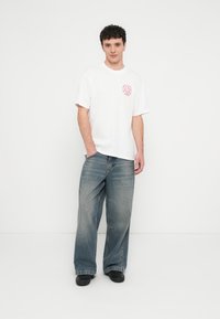 Bela bombažna majica z rdečim grafičnim tiskom na prsih, kombinirana z ohlapnimi, izpiranimi modrimi jeans hlačami in črno obutvijo.