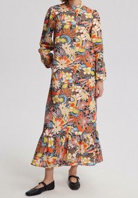 Femme portant une robe florale à manches longues et longueur cheville avec un ourlet volants et des chaussures Mary Jane noires, se tenant devant un fond uni.