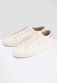 Sneakers beige in tela con suole in gomma bianche, punta rotonda, design con lacci e un pannello laterale testurizzato come accento. Stile semplice e funzionale.