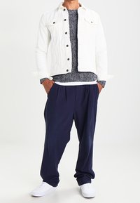 Weiße Jeansjacke, strukturierter marineblauer Pullover, locker sitzende dunkle Hose, weiße Sneakers. Kontrast von Materialien und Farben in einem lässigen Outfit.
