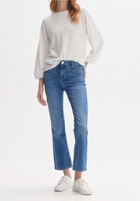 Haut à manches longues texturé blanc associé à un jean court bleu clair. Des baskets blanches complètent la tenue. Fond uni.