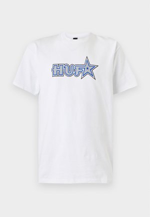 Jeune homme aux cheveux courts et bouclés porte un T-shirt blanc avec un texte bleu "HUF" et une étoile, ainsi qu'un pantalon noir avec de petites pièces orange et bleues.