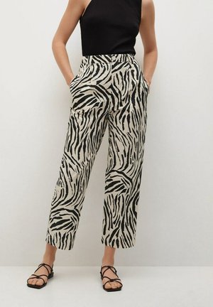 Frau trägt hoch taillierte, weit geschnittene Zebra-Print-Hosen, schwarzes ärmelloses Oberteil und schwarze Sandalen mit Riemchen, steht mit den Händen in den Taschen.