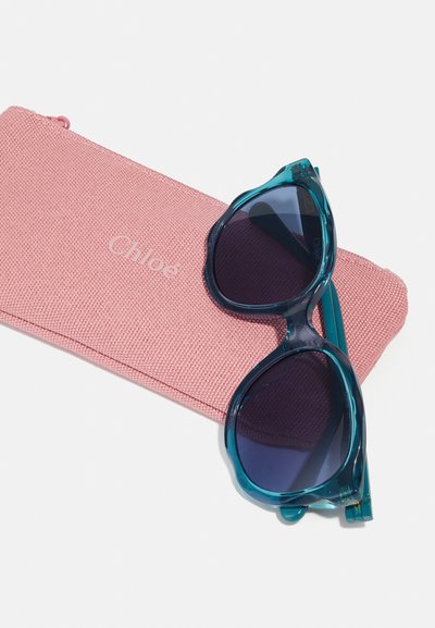 Occhiali da sole turchesi con lenti scure, appoggiati su una custodia rosa testurizzata con la scritta "Chloé" in rilievo. La custodia presenta una chiusura con zip.