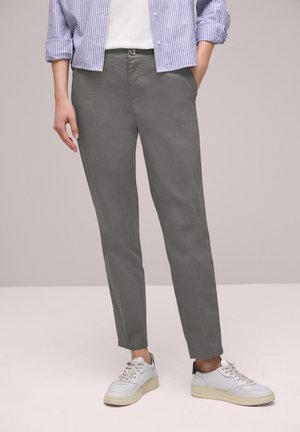 Persoon draagt grijze rechte broek, witte sneakers en een blauw-wit gestreept shirt met opgerolde mouwen, staat met één hand in de zak.