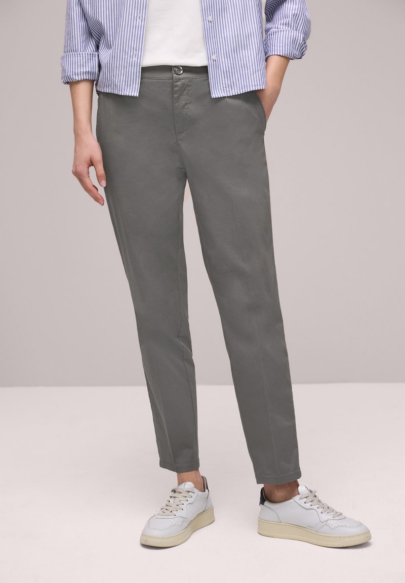 Person trägt graue gerade geschnittene Hose, weiße Sneakers und ein blau-weiß gestreiftes Hemd mit hochgekrempelten Ärmeln, steht mit einer Hand in der Tasche.