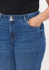 Jeans din denim albastru, cu talie înaltă, cu o textură netedă, două butoane metalice și buzunare laterale. Prezintă o cusătură subtilă de-a lungul cusăturilor.