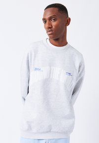 Sudadera gris con cuello y puños acanalados, que presenta un gran texto blanco "AROUND THE WORLD" y un pequeño detalle de etiqueta azul en el pecho.