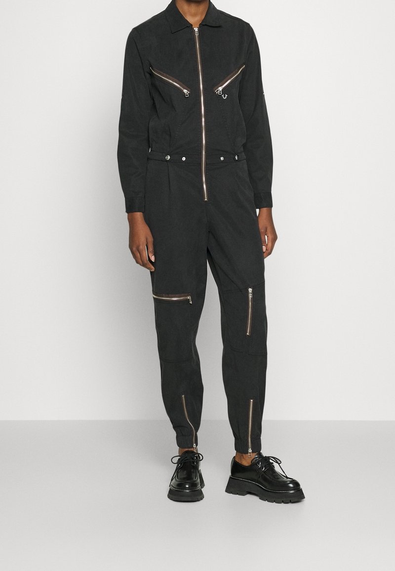Personne portant une combinaison noire à manches longues avec plusieurs fermetures éclair sur le devant et des chaussures noires épaisses à lacets, debout devant un fond clair uni.