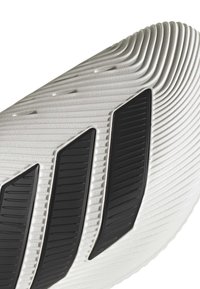 Zbliżenie na teksturowaną, białą podeszwę trampka z trzema czarnymi, ukośnymi paskami i żłobionymi wzorami.