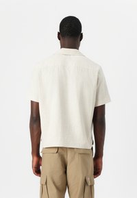 Camisa beige de manga corta con un patrón texturizado, que cuenta con un cuello y un corte relajado, combinada con pantalones cargo caqui.