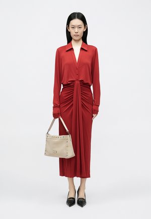 Robe rouge à manches longues avec un nœud sur le devant et une fermeture à boutons. Accessoirisée d'un sac à main beige orné de clous décoratifs et de talons pointus noirs.