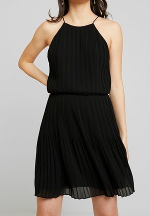 Frau trägt ärmelloses schwarzes, plissiertes Kleid mit Spaghettiträgern, steht mit einer Hand in der Hüfte vor neutralem Hintergrund.