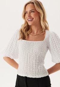 Blusa bianca con texture, scollo quadrato e maniche a sbuffo a metà. Presenta un tessuto rugoso e una vita aderente, abbinata a pantaloni neri.