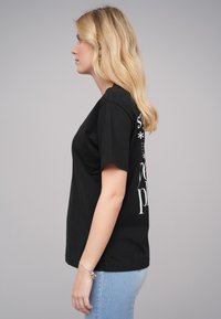 T-shirt en coton noir avec col rond et manches courtes. Design en texte blanc au dos. Porté avec un jean bleu clair.
