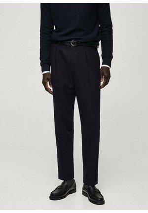 Homme portant un pantalon habillé noir, une ceinture noire, un pull noir et des chaussures en cuir noires, debout devant un fond clair uni.