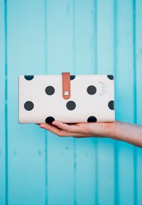 White wallet with large black polka dots, featuring a brown strap with a snap closure and a silver accent, held against a blue background.
