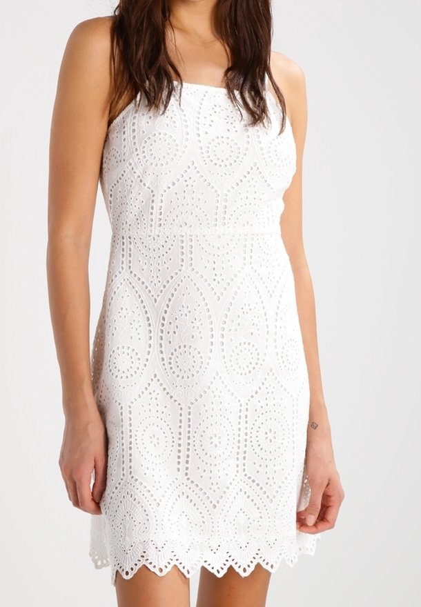 Femme portant une robe blanche sans manches avec des motifs complexes circulaires et ovales ajourés et un ourlet festonné.