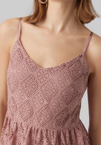 Vestido de encaje rosa con escote en V, tirantes finos y dobladillo acampanado. La tela tiene un intrincado patrón de recortes y una textura suave.