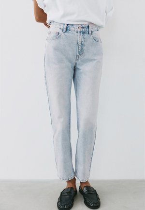Persoon draagt lichtblauwe high-waisted jeans en zwarte slip-on loafers, staand tegen een effen witte achtergrond.