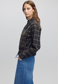 Camicia a quadri marrone con design ricamato floreale, abbinata a una lunga gonna di jeans. Presenta una vita con coulisse e tasche.