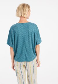 Žalios spalvos trumpas palaidinė su zigzago raštu, pasižyminti laisvu siluetu ir lygiu paviršiumi. Kombinuojama su juostuotomis, plačiomis kelnėmis šviesių atspalvių.