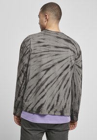 Majica s dugim rukavima u tie-dye uzorku sive boje s crnim spiralnim uzorkom. Mehak pamučni materijal s ležernim krojem i okruglim izrezom. Nosiva preko ljubičaste majice.