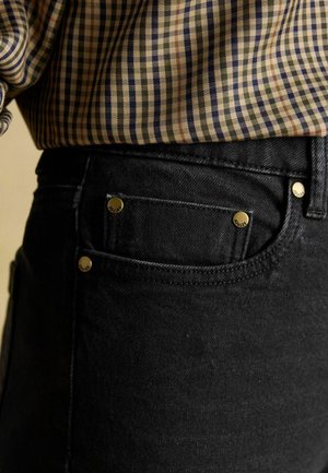 Schwarze Jeans aus Denim mit goldenen Akzenten, ausgestattet mit einer Vordertasche und einem strukturierten Stoff. Kombiniert mit einem gemusterten, karierte Hemd.