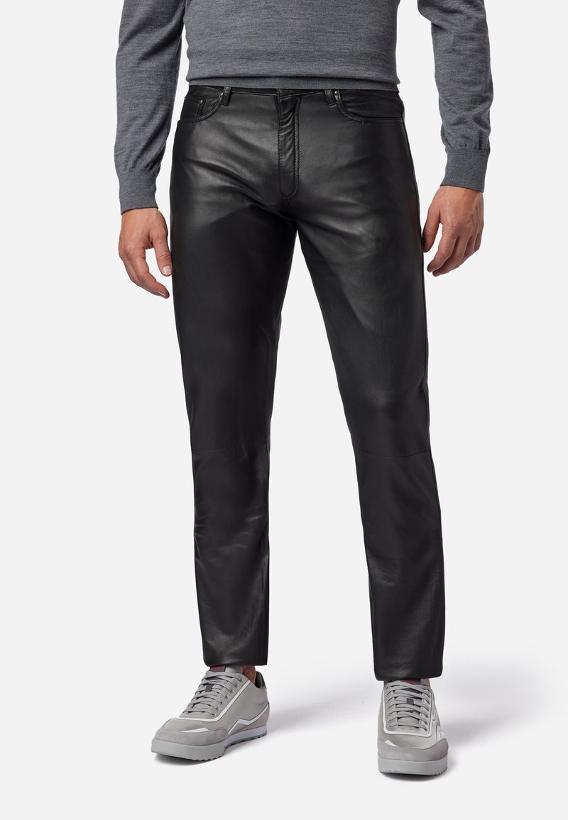 Zwarte leren broek met een slim fit ontwerp, gekenmerkt door een gladde textuur en zichtbare stiksels. Gecombineerd met grijze sneakers, wat een casual uitstraling geeft.