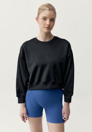 Mladá žena s blond vlasmi má na sebe čiernu oversize mikinu a priliehavé modré rebrované cyklistické šortky na jednoduchom svetlom pozadí.