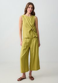 Top senza maniche verde oliva con dettaglio anteriore attorcigliato e accentuazione metallica, abbinato a pantaloni a gamba larga coordinati. Sandali piatti marroni.