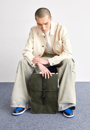 Zaino in pelle verde con pattina, tracolla regolabile e fibbia nera; modello seduto in giacca chiara e jeans larghi, sneakers con dettagli blu.