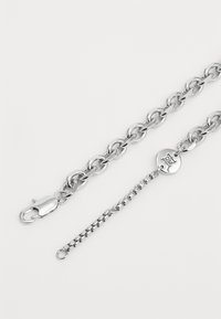 Silver metal chain bracelet with large oval links, featuring a lobster clasp and an adjustable extension chain. Circular logo tag present.
