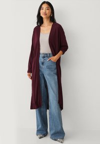Bordeauxkleurige cardigan met open voorkant, lange mouwen, gedragen over een licht tanktop, gecombineerd met hooggevNew jeans met wijde pijpen en gestreepte schoenen.