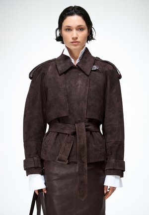 Žena s krátkymi tmavými vlasmi, oblečená v tmavohnedej semišovej bunde s opaskom, cez koženú sukňu a bielu košeľu s predĺženými manžetami.