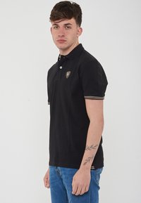 Polo shirt nero con maniche corte, dotato di colletto con bottoni, logo dorato sul petto e dettagli a righe sulle maniche.