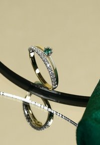 Gold- und Silberring mit geschwungenem Design, ausgestattet mit einem grünen Edelstein und einer Reihe weißer Steine entlang des Rings, die auf einer Oberfläche reflektieren.