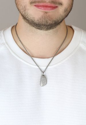 Silberne Kettenfassung mit einem strukturierten, rechteckigen Anhänger mit punktierten Mustern. Trägt man über einem weißen, strukturierten Hemd.