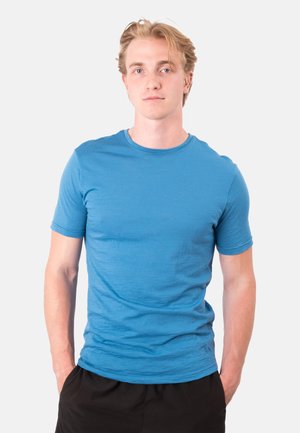 Junger Mann mit blondem Haar, der ein blaues T-Shirt und schwarze Hose trägt, steht mit den Händen in den Taschen vor einem einfarbigen Hintergrund.