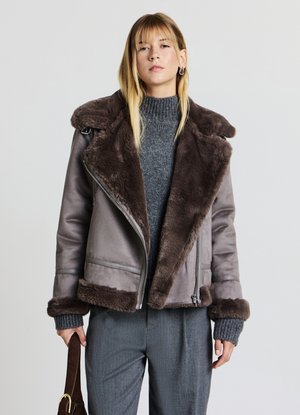 Graue Bikerjacke mit braunem Kunstfellkragen und -manschetten, mit Reißverschluss und glatter Textur, kombiniert mit einem grauen Rollkragenpullover und Hose.