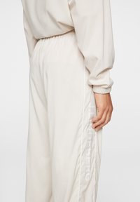 Beige joggers med elastisk midjeband, vit kant längs sidorna och en tryckt logotyp i vitt på vänster ben. Slät, lätt tyg.